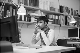 A woman seated at a table in front of a monitor with a lamp above, holds her hands together and rests her lips against her fingers. Shelves of folders are seen above her. Another identical workstation with a lamp above, behind her.