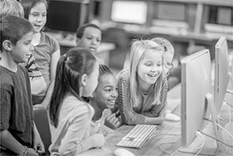 A group of children are crowded in front of two monitors on a table in a classroom. The three girls in front are smiling as they look at the screen.